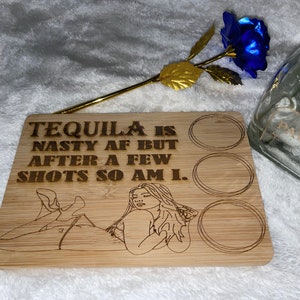Könnte beinhalten: Ein Holztablett mit drei Halterungen für Schnapsgläser. Das Tablett ist mit einem gravierten Design einer Frau und dem Text "TEQUILA IS NASTY AF BUT AFTER A FEW SHOTS SO AM I." verziert.