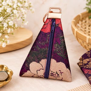 May include: A triangular coin pouch featuring a floral pattern in shades of deep purple, red, and beige. The pouch has a dark blue vertical zipper closure, a rose gold metal hardware top attachment, and a matching circular metal key ring.