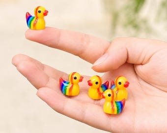 May include: A collection of small, yellow duck figurines with rainbow-colored wings and accents. The ducks have red beaks and are held in a hand. The ducks are a cheerful and colorful decorative item.