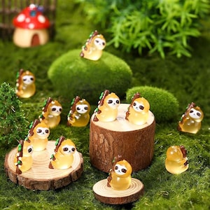 May include: A collection of miniature sloth figurines in various poses, crafted from translucent yellow resin. Each sloth has a white face, black eyes, and a brown branch detail. The figurines are arranged on a bed of green moss and wooden platforms.