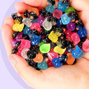 May include: A handful of colorful, miniature penguin figurines. Each penguin is a different translucent color, including blue, pink, yellow, and green, and wears a small black hat. The penguins are held in an open hand.