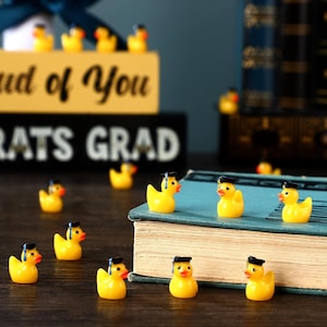 May include: A collection of small, yellow rubber duck figurines, each wearing a tiny graduation cap. The ducks are scattered around a book and a sign that reads "Congrats Grad" in black and gold. The scene suggests a graduation theme.