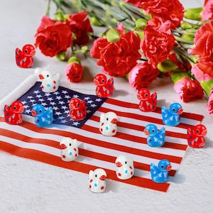May include: A collection of small, colorful duck figurines in red, white, and blue, arranged on an American flag. The ducks have star patterns and the word "USA" on them. Red carnations are in the background.