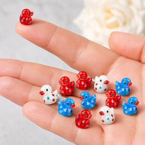 May include: A collection of miniature duck figurines in red, white, and blue, decorated with stars. One duck sits on a fingertip. The ducks have a glossy finish and are arranged in a hand.