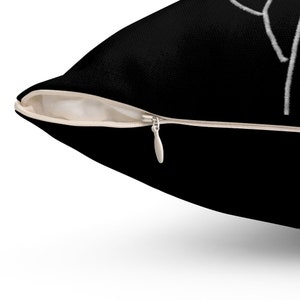 May include: Black throw pillow with a white zipper and a white abstract design. The pillow has a black fabric exterior and a beige interior. The zipper is silver.