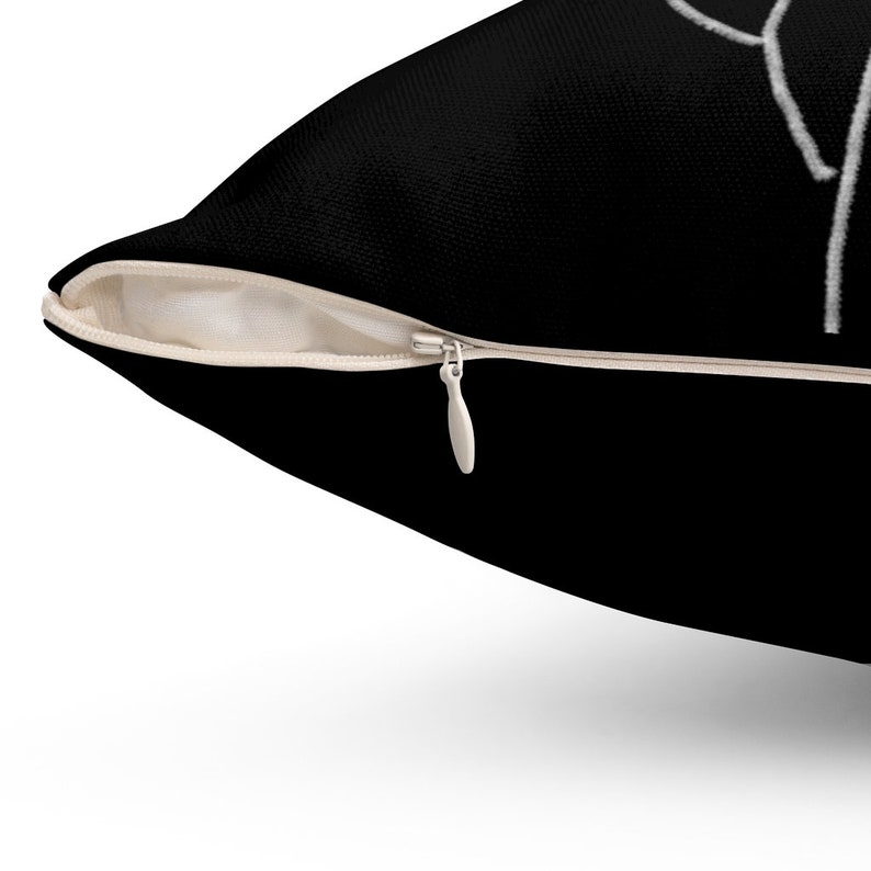 May include: Black throw pillow with a white zipper and a white abstract design. The pillow has a black fabric exterior and a beige interior. The zipper is silver.