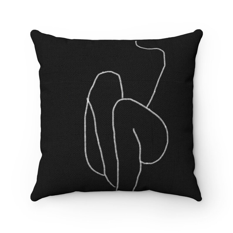 May include: Square black throw pillow featuring a minimalist white line drawing of a human figure. The pillow has a textured appearance, with the white lines creating a simple, elegant design. The pillow is square and appears to be made of fabric.