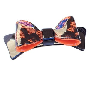 May include: A black and orange bow hair clip with a lace pattern. The bow is made of plastic and has a shiny finish.