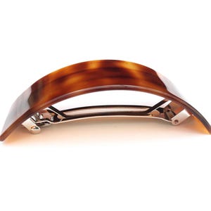 Könnte beinhalten: Eine Haarklammer mit Schildpattmuster und geschwungenem Design. Die Spange hat eine glänzende, braune und bernsteinfarbene Oberfläche und einen silberfarbenen Metallverschluss. Dieses Haaraccessoire ist zum Halten der Haare konzipiert.