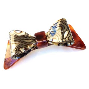 May include: A hair clip featuring a bow design with a marbled pattern in shades of brown, cream, and black. The center of the bow is secured by a solid dark brown band, and the base of the clip is a translucent amber-colored triangular shape.