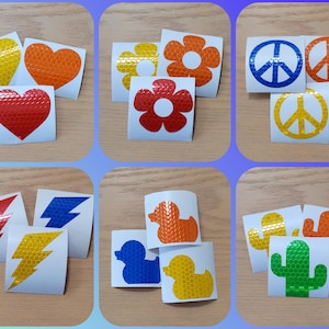 May include: Assortment of reflective stickers in various shapes and colors, including hearts, flowers, lightning bolts, peace signs, ducks, and cacti. The stickers are all made of a reflective material and have a textured surface.