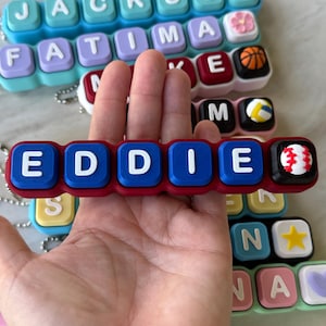 May include: A rectangular silicone keychain featuring the name EDDIE in white block letters on blue square segments, with a red base and a baseball charm at the end. Other similar keychains with various names and charms are visible in the background.