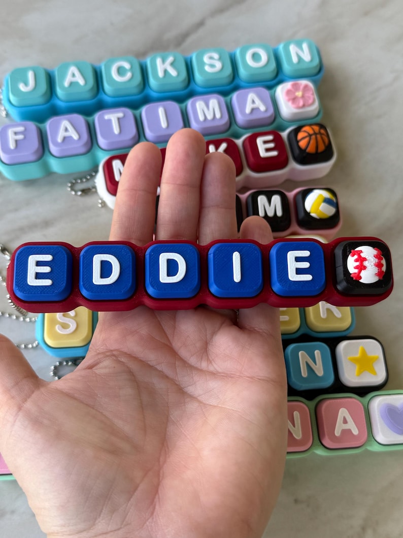 May include: A rectangular silicone keychain featuring the name EDDIE in white block letters on blue square segments, with a red base and a baseball charm at the end. Other similar keychains with various names and charms are visible in the background.