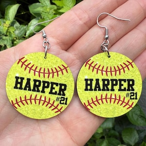 May include: A pair of softball-themed earrings. Each earring is a yellow circle with red stitching and the name "HARPER" in black block letters, with "#21" below. The earrings have silver-colored fish hook ear wires.