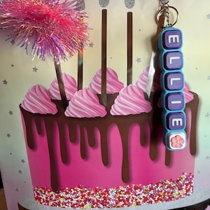 May include: A personalized keychain featuring rectangular blue and purple beads spelling the name ELLIE, topped with a small pink flower charm. The keychain is attached to a metal ring and clasp, displayed against a birthday gift bag with a cake graphic.