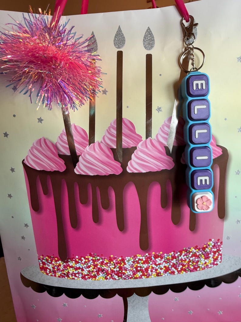 May include: A personalized keychain featuring rectangular blue and purple beads spelling the name ELLIE, topped with a small pink flower charm. The keychain is attached to a metal ring and clasp, displayed against a birthday gift bag with a cake graphic.