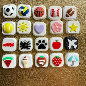 May include: A collection of twenty square white tiles, each featuring a distinct raised icon. The icons include various sports balls, a sun, a heart, a flower, a star, a car, a spider, a paw print, an apple, an airplane, a watermelon slice, an ice cream cone, strawberries, and an umbrella.