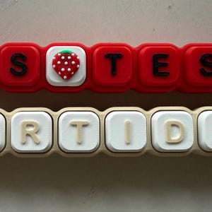 May include: Two rectangular silicone keychains with metal ball chains. The top red keychain features black letters spelling MS TESS with a strawberry icon. The bottom beige keychain features white letters spelling PARTIDA with a coffee cup icon.