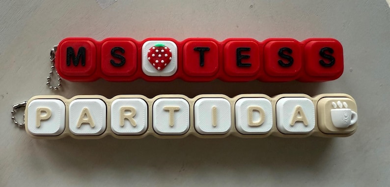 May include: Two rectangular silicone keychains with metal ball chains. The top red keychain features black letters spelling MS TESS with a strawberry icon. The bottom beige keychain features white letters spelling PARTIDA with a coffee cup icon.