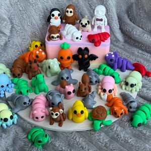 May include: A collection of colorful, miniature clay figurines in various animal and creature shapes, including a penguin, monkey, pug, and octopus. The figurines are arranged on a white circular base and a pink box.