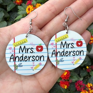 May include: A pair of round wooden earrings with a white background and blue lines. The earrings feature a ruler, scissors, a pencil, hearts, and a red apple with a white heart. The text "Mrs. Anderson" is written in black on each earring.
