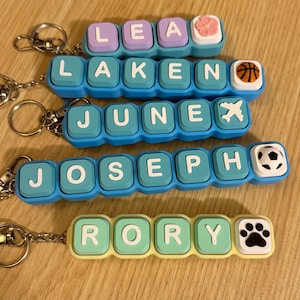 May include: Four custom keychains featuring square, button-like blocks in pastel blue, teal, and light green. Each keychain displays a name: LEA with a flower, LAKEN with a basketball, JUNE with an airplane, JOSEPH with a soccer ball, and RORY with a paw print.