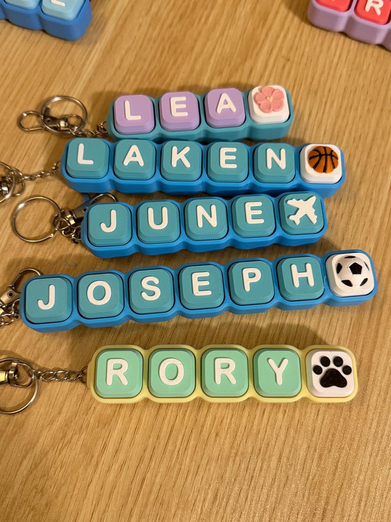May include: Four custom keychains featuring square, button-like blocks in pastel blue, teal, and light green. Each keychain displays a name: LEA with a flower, LAKEN with a basketball, JUNE with an airplane, JOSEPH with a soccer ball, and RORY with a paw print.