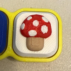 May include: A square-shaped button with a bright yellow border and a white center. Centered on the white surface is a three-dimensional mushroom with a red cap featuring white spots and a light brown stem, all showing a textured 3D printed finish.