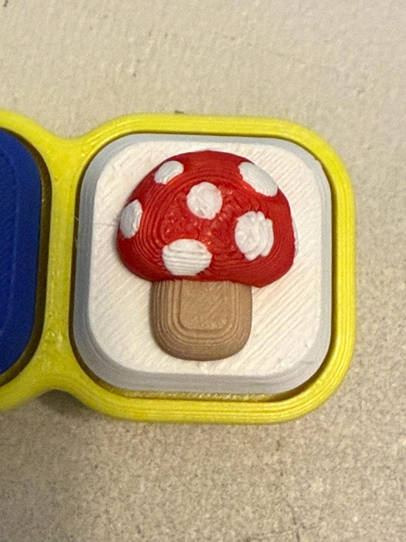 May include: A square-shaped button with a bright yellow border and a white center. Centered on the white surface is a three-dimensional mushroom with a red cap featuring white spots and a light brown stem, all showing a textured 3D printed finish.
