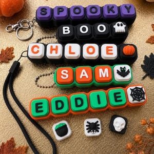 May include: A collection of silicone keychains featuring block letters spelling SPOOKY, BOO, CHLOE, SAM, and EDDIE. The keychains include colorful charms shaped like a ghost, pumpkin, bat, and spiderweb, along with loose charms of a cauldron, spider, and skull.