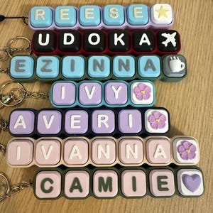 May include: A collection of rectangular keychains featuring raised, square-shaped letter tiles in various pastel and dark colors. Each keychain displays a name, including REESE, UDOKA, EZINNA, IVY, AVERI, IVANNA, and CAMIE, with decorative icons.