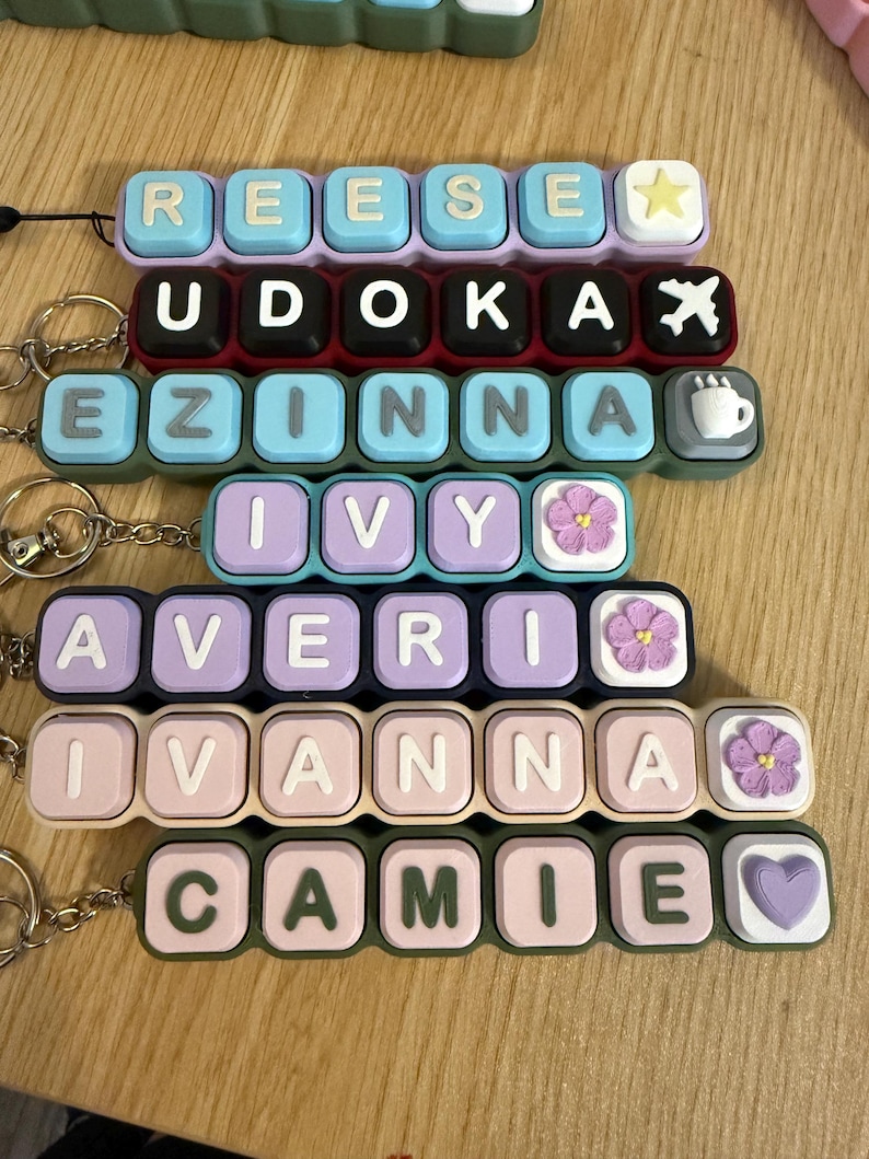 May include: A collection of rectangular keychains featuring raised, square-shaped letter tiles in various pastel and dark colors. Each keychain displays a name, including REESE, UDOKA, EZINNA, IVY, AVERI, IVANNA, and CAMIE, with decorative icons.