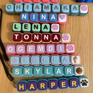 May include: A collection of eight rectangular silicone keychains, each featuring colorful square buttons with letters spelling out the names ERICA, CHIAMAKA, NINA, LENA, TONNA, OGEMDI, STELLA, SKYLAR, and HARPER, accented with small decorative charms.