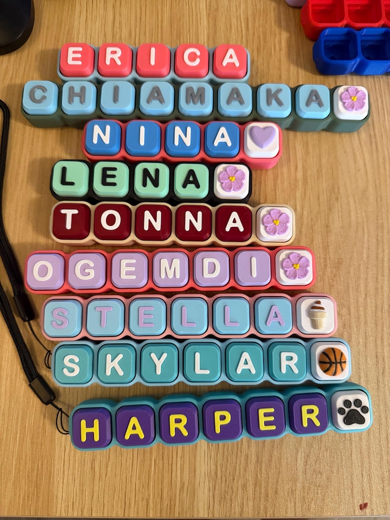 May include: A collection of eight rectangular silicone keychains, each featuring colorful square buttons with letters spelling out the names ERICA, CHIAMAKA, NINA, LENA, TONNA, OGEMDI, STELLA, SKYLAR, and HARPER, accented with small decorative charms.