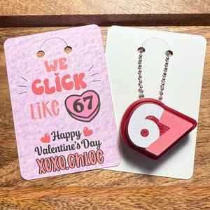 May include: A Valentine's Day themed gift set featuring a red and white pendant with the number 67, hanging from a silver chain, and a pink card with the text "We Click Like 67 Happy Valentine's Day XOXO.Chloe".
