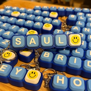 May include: A custom keychain featuring light blue square blocks with white letters spelling SAUL, followed by a white block with a yellow smiley face. The keychain is constructed from multiple connected plastic segments with a metal ring attached.