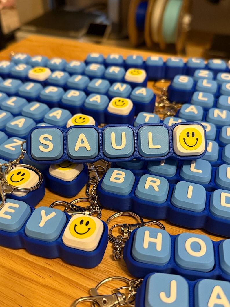 May include: A custom keychain featuring light blue square blocks with white letters spelling SAUL, followed by a white block with a yellow smiley face. The keychain is constructed from multiple connected plastic segments with a metal ring attached.