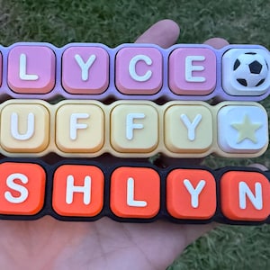 May include: Three horizontal rows of rectangular silicone charms featuring raised white letters and icons. The top row is lavender with ELYCE and a soccer ball. The middle row is yellow with PUFFY and a star. The bottom row is black with ASHLYN and a softball.