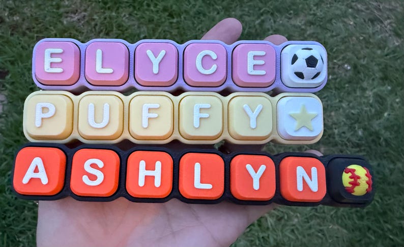 May include: Three horizontal rows of rectangular silicone charms featuring raised white letters and icons. The top row is lavender with ELYCE and a soccer ball. The middle row is yellow with PUFFY and a star. The bottom row is black with ASHLYN and a softball.