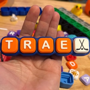 May include: A blue plastic keychain featuring five square tiles with rounded corners. Four orange tiles display the white letters T, R, A, and E, while the fifth white tile features a black graphic of two crossed hockey sticks and a puck.