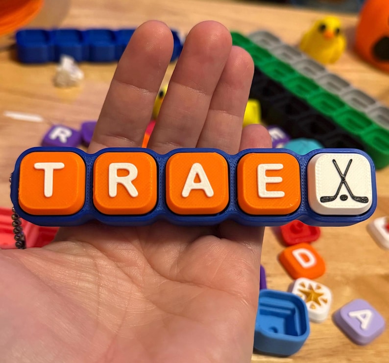 May include: A blue plastic keychain featuring five square tiles with rounded corners. Four orange tiles display the white letters T, R, A, and E, while the fifth white tile features a black graphic of two crossed hockey sticks and a puck.
