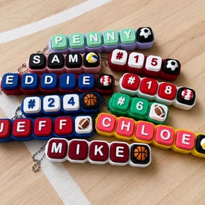May include: A collection of colorful name tags with sports-themed icons. Each tag features a name or number spelled out in white letters on a colored background, with sports balls like soccer, volleyball, basketball, and baseball.