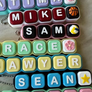 May include: A collection of personalized keychains featuring square, colorful plastic blocks with raised white letters spelling names like Jackson, Fatima, Mike, Sam, Grace, Sawyer, Sean, and Katrina, accented with small icons like flowers, balls, and stars.