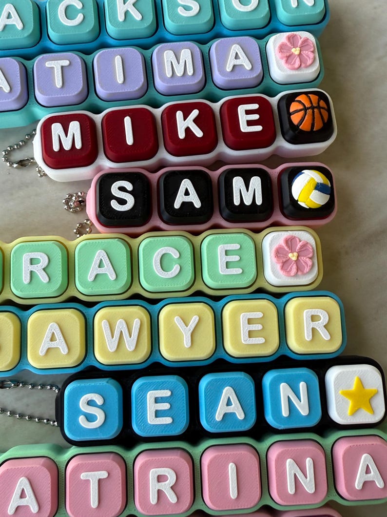 May include: A collection of personalized keychains featuring square, colorful plastic blocks with raised white letters spelling names like Jackson, Fatima, Mike, Sam, Grace, Sawyer, Sean, and Katrina, accented with small icons like flowers, balls, and stars.