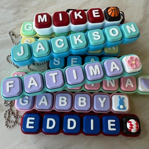 May include: A collection of personalized silicone keychains featuring raised white letters on colorful square blocks. Names include MIKE with a basketball, JACKSON, FATIMA with a pink flower, GABBY with a blue bunny, and EDDIE with a baseball.