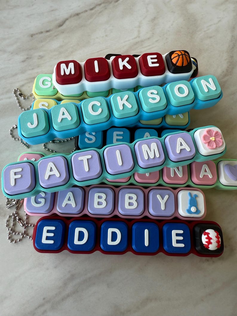 May include: A collection of personalized silicone keychains featuring raised white letters on colorful square blocks. Names include MIKE with a basketball, JACKSON, FATIMA with a pink flower, GABBY with a blue bunny, and EDDIE with a baseball.