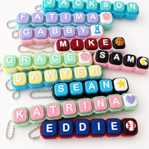 May include: A collection of personalized keychains featuring colorful, square, keyboard-style keys with white letters spelling names including Jackson, Fatima, Gabby, Mike, Grace, Sam, Sawyer, Sean, Katrina, and Eddie, accented with small icons.