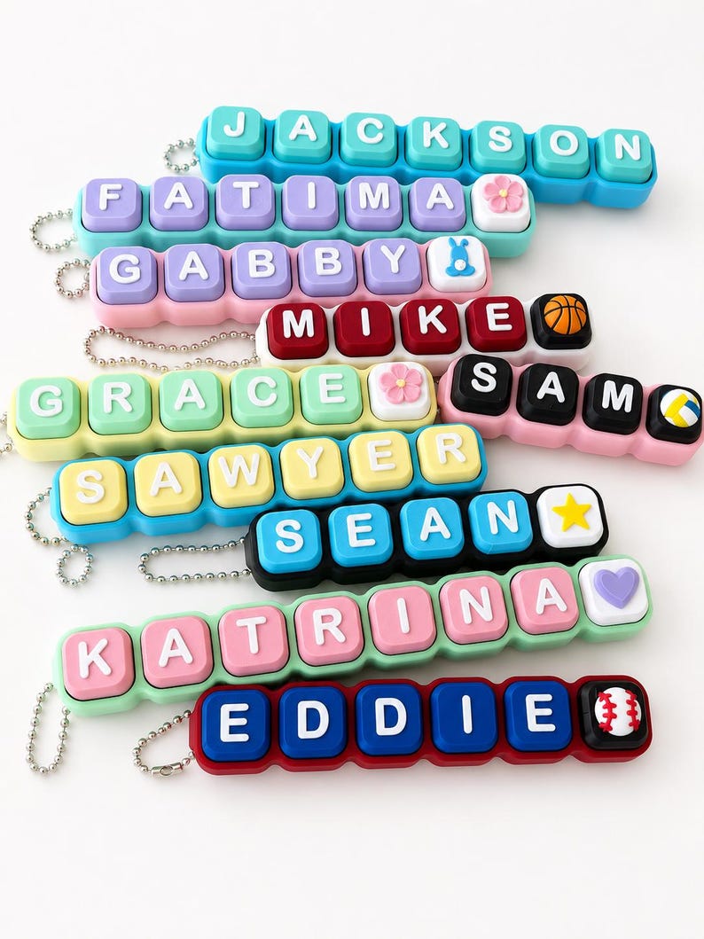 May include: A collection of personalized keychains featuring colorful, square, keyboard-style keys with white letters spelling names including Jackson, Fatima, Gabby, Mike, Grace, Sam, Sawyer, Sean, Katrina, and Eddie, accented with small icons.