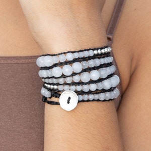 K&ouml;nnte beinhalten: Ein Stapel wei&szlig;er Edelstein-Armb&auml;nder mit schwarzem Band und einem silbernen Anh&auml;nger. Die Armb&auml;nder sind &uuml;bereinander am Handgelenk einer Person gelegt.