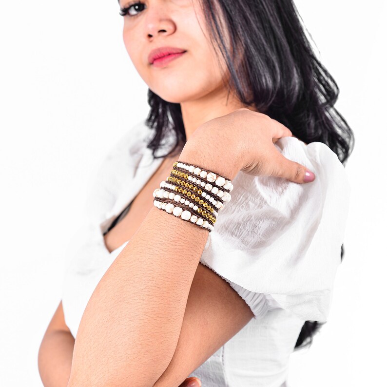 May include: A close-up of a woman's arm wearing a multi-layered bracelet with brown leather cord, white beads, and gold accents.